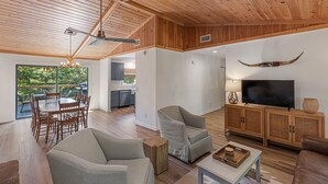 A spacious living area with a wooden ceiling, a dining set, a TV on a wooden cabinet, and a large window overlooking greenery. - Private Renovated home 1.5 Miles to downtown Highlands!  (Highlands)