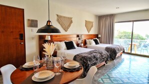 Une chambre d’hôtel avec une tête de lit en bois, deux lits, un coin repas avec une table dressée pour quatre personnes, et une grande fenêtre donnant sur l’extérieur.