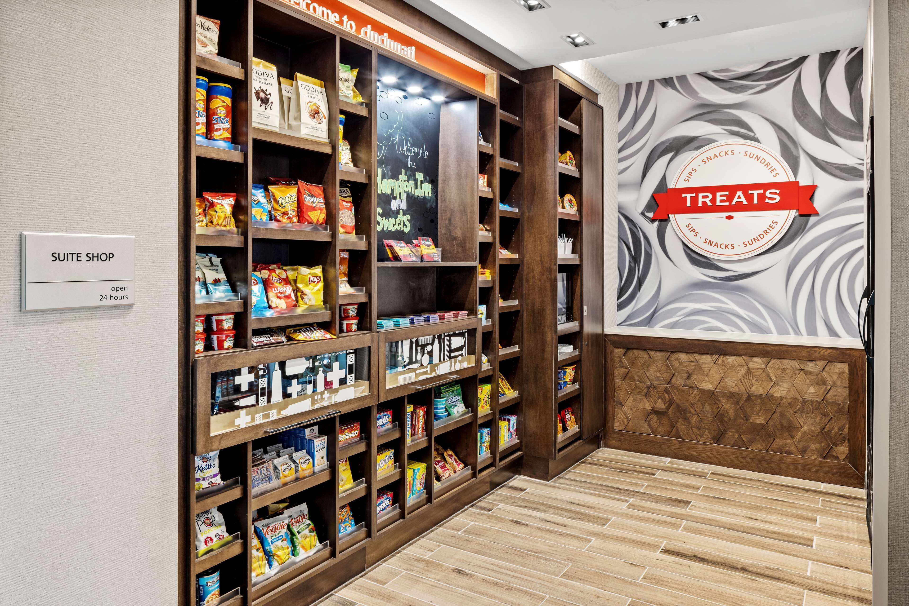 A suite shop with shelves stocked with snacks and sweets, a sign indicating it's open 24 hours, and a wall with a 'Treats' sign.