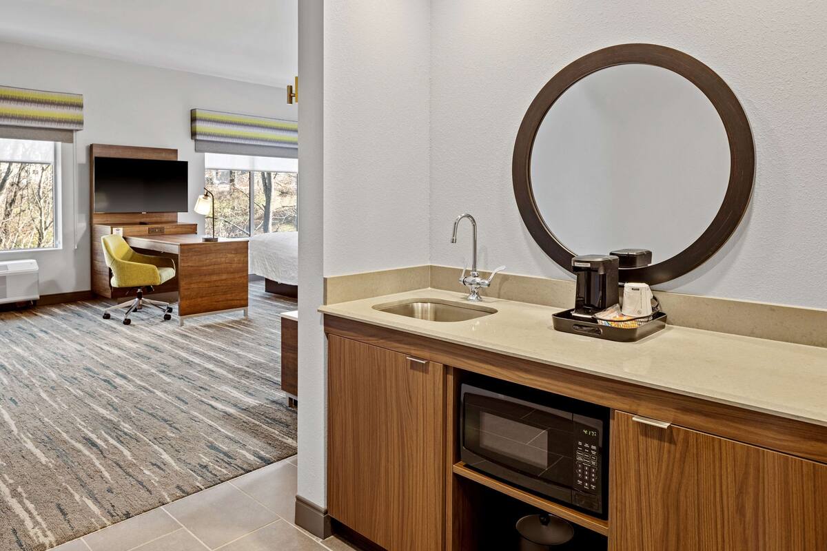 A hotel room with a wooden desk, a yellow chair, a large mirror, a sink, and a microwave.