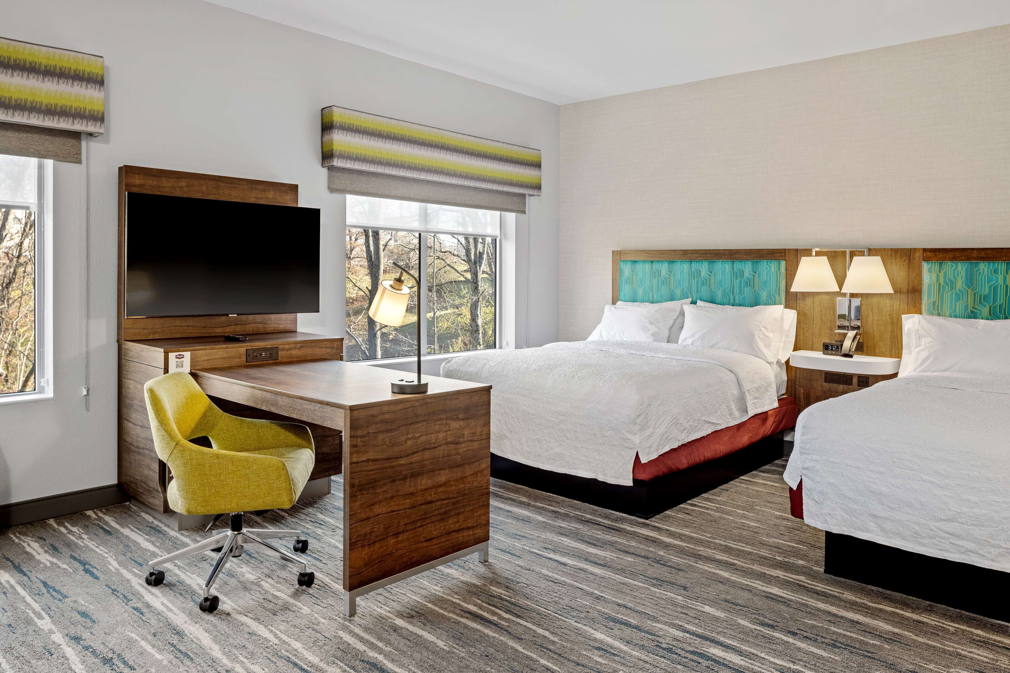 A hotel room with a large flat-screen TV, two beds, a desk with a chair, and a view of trees through the window.