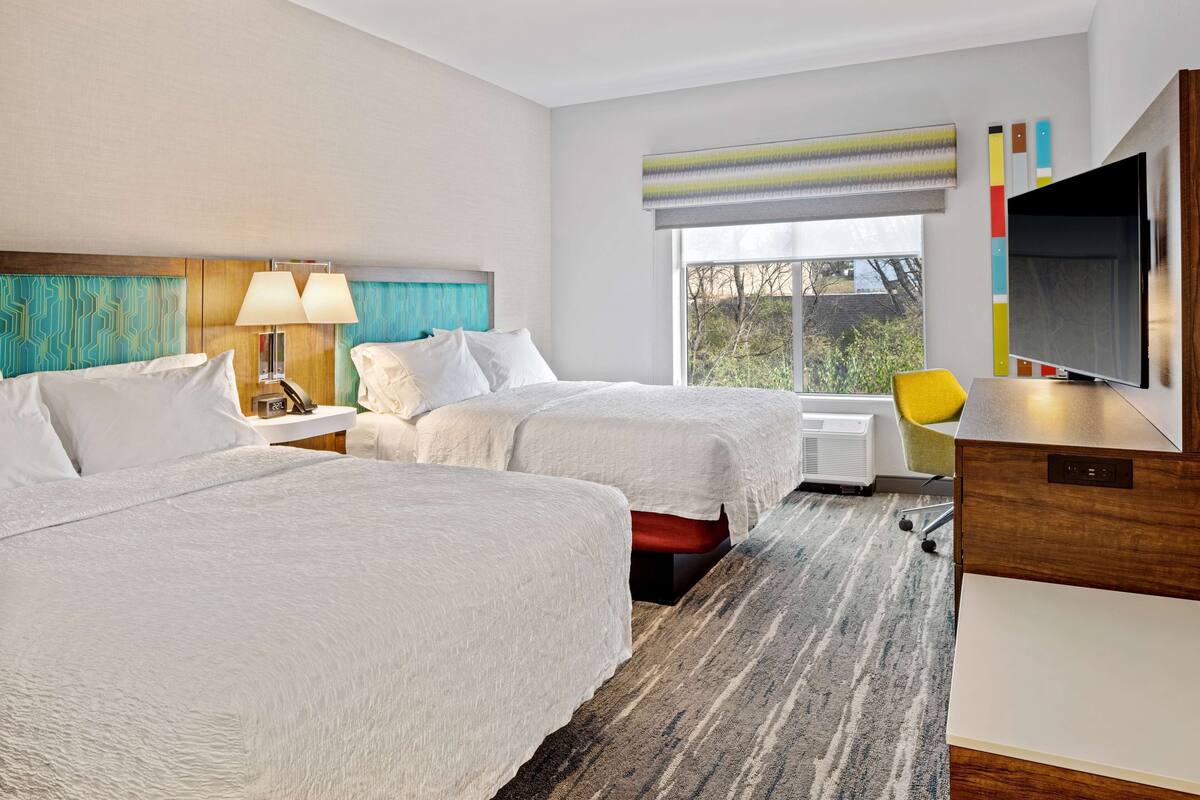 A hotel room with two beds, a desk, a TV, and a window with a view.