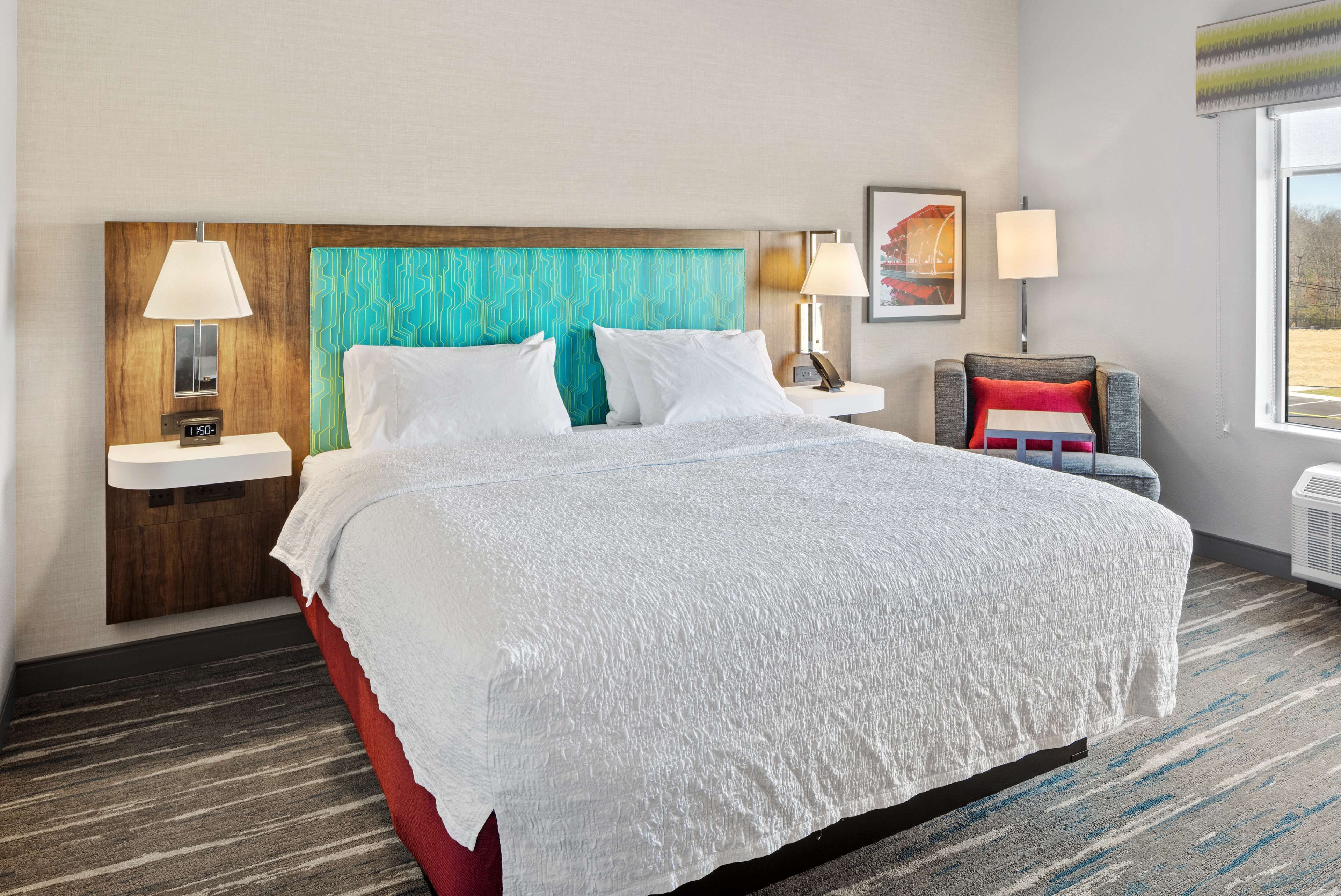 Habitación de hotel con una cama grande, cabecera de madera, mesita de noche con lámpara, una silla con cojín rojo y ventana con vistas.