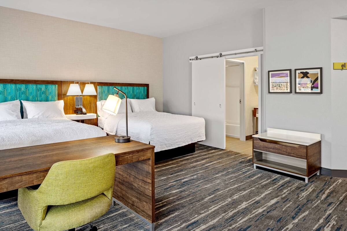 A hotel room with two beds, a desk, a chair, a bathroom with a sink, and a door leading to another room.