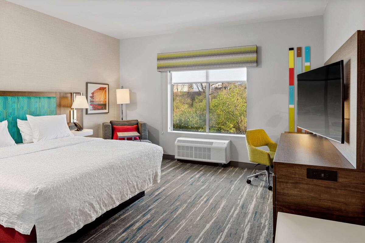 A hotel room with a large bed, a desk, a chair, a TV, and a window with a view of trees.