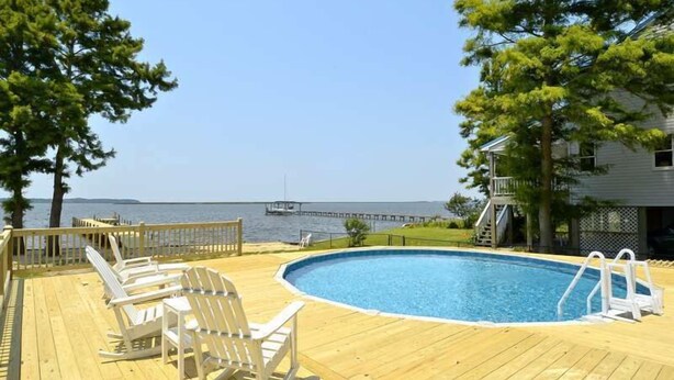 Um deck de madeira com piscina, espreguiçadeiras e vista para a água.