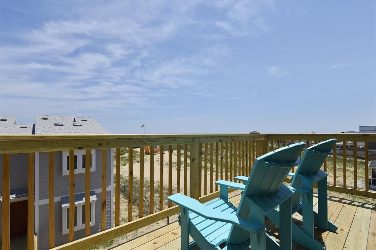 A wooden deck with two blue Adirondack chairs, overlooking a beach with a clear sky.