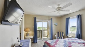 Habitación de hotel con cama, televisión montada en la pared, ventilador de techo, escritorio con lámpara, una silla y vista a la playa por la ventana.