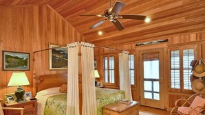 A four-poster bed with a canopy, wooden walls, a ceiling fan, and a wicker chair. - Gorgeous old nags head oceanfront home with spacious decks and beautiful views! (Nags Head)