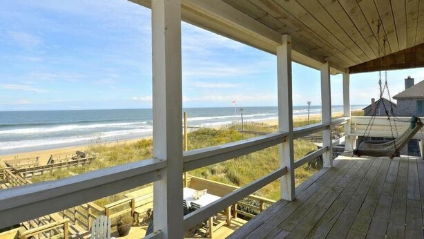 A wooden deck overlooking a beach with a view of the ocean and a flag in the distance. - Gorgeous old nags head oceanfront home with spacious decks and beautiful views! (Nags Head)