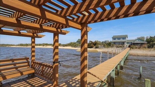 A wooden deck with a pergola structure, a wooden bench, and a view of a house across a body of water. - Cozy sound front cottage in Nags Head! Incredible views and right next to JRSP! (Nags Head)