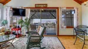 A cozy living room with a television, wicker chairs, a glass table, and a large sliding glass door leading to an outdoor area. - Cozy sound front cottage in Nags Head! Incredible views and right next to JRSP! (Nags Head)