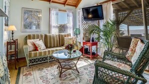 A cozy living room with a sofa, a glass table, wicker chairs, a TV, and a large window with curtains. - Cozy sound front cottage in Nags Head! Incredible views and right next to JRSP! (Nags Head)