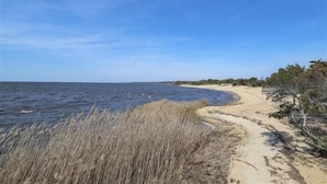 A beach with a sandy path leading to the water, surrounded by dry grass and sparse vegetation. - Cozy sound front cottage in Nags Head! Incredible views and right next to JRSP! (Nags Head)