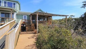 A wooden deck leading to a covered porch with a view of a house and surrounding vegetation. - Cozy sound front cottage in Nags Head! Incredible views and right next to JRSP! (Nags Head)