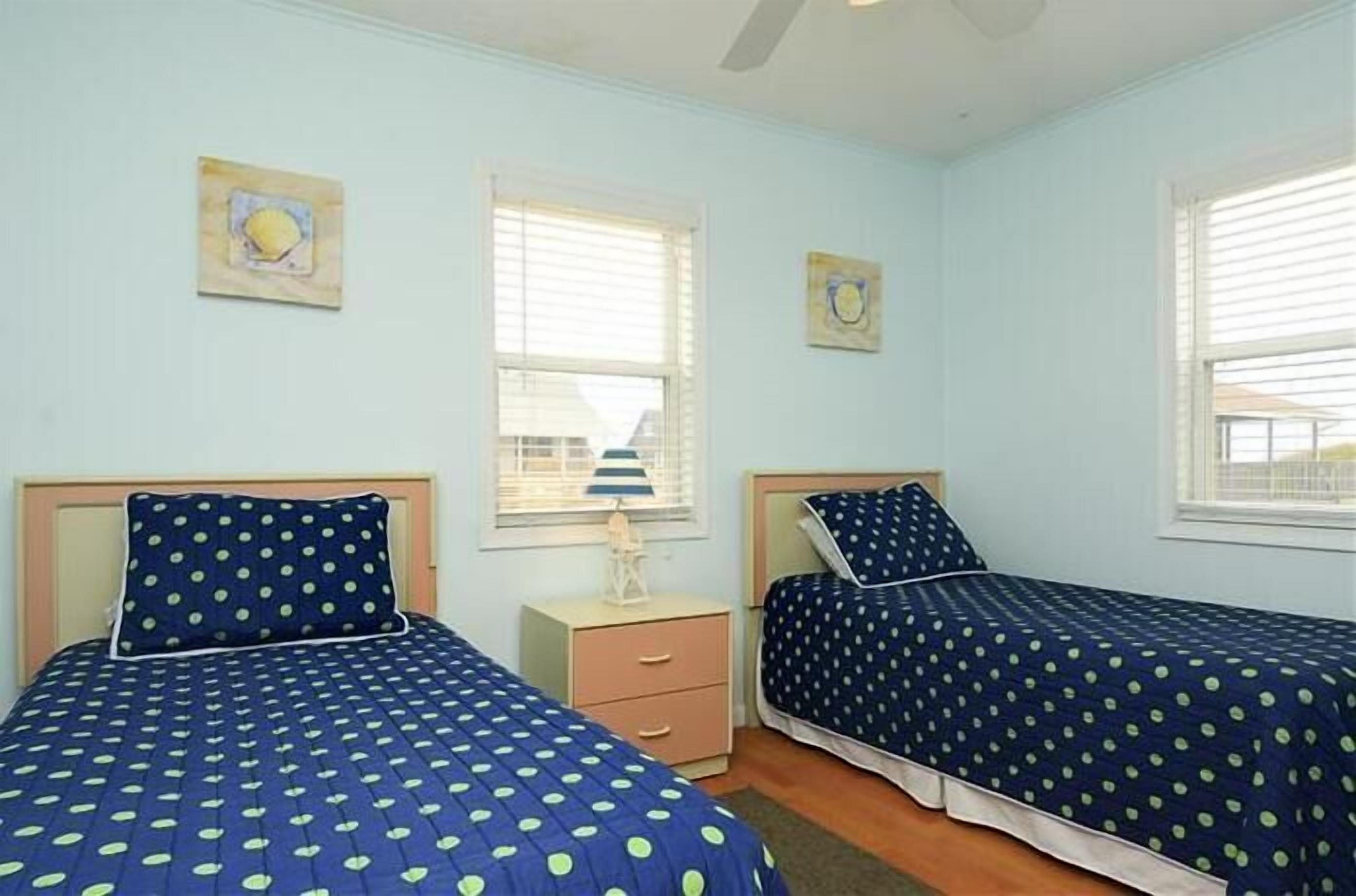 A room with two beds, each with a blue polka dot bedspread, a small bedside table, and a window with blinds.