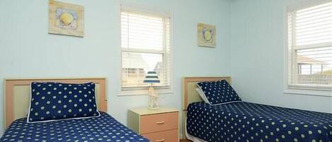 A room with two beds, each with a blue polka dot bedspread, a small bedside table, and a window with blinds.