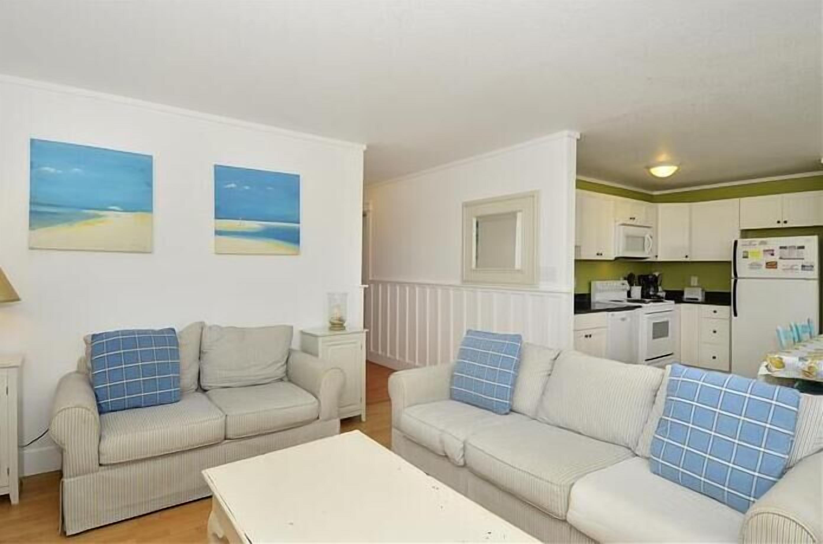 A compact living space with two sofas and a dining table, a kitchenette with a microwave and refrigerator, and two beach-themed wall art pieces.