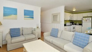 Ein kompakter Wohnbereich mit zwei Sofas und einem Esstisch, eine Küchenzeile mit Mikrowelle und Kühlschrank, sowie zwei Wandbilder im Strandhausstil.
