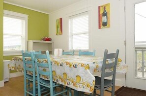 A dining area with a table covered in a floral tablecloth, surrounded by blue chairs. There are two windows with blinds, a shelf with a red object, and a door with a doormat.