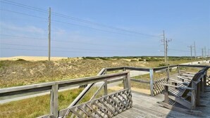 Ein hölzerner Gehweg mit Metallgeländern führt zu einem Sandstrandabschnitt mit Dünen und Stromleitungen im Hintergrund.