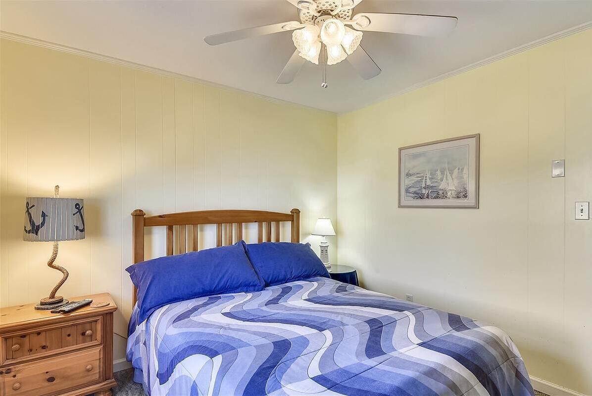 Une chambre à coucher avec un lit, une table de chevet en bois, deux lampes, un ventilateur de plafond et un tableau encadré au mur.