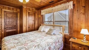 A wooden cabin bedroom with a bed, a nightstand, a lamp, and a window with blinds. - Beautiful and classic OBX oceanfront home in KDH! Steps from the Avalon Pier. (Kill Devil Hills)