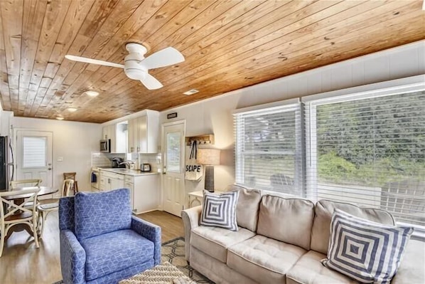 A modern living room with a wooden ceiling, a blue armchair, a beige sofa with patterned pillows, and a view of the outdoors through large windows.