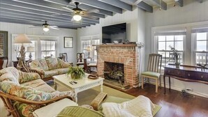 A spacious living room with a brick fireplace, wicker furniture, and a ceiling fan. - 6 bedroom, classic old nags head style oceanfront with a hot tub! (Nags Head)