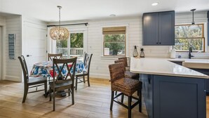 A modern kitchen with dark cabinets, a large island, and a dining area with wicker chairs and a table. - Private pool and steps to the beach in quiet Southern Shores! Perfect beach home (Southern Shores)