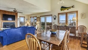 A spacious living room with a large blue sofa, wooden dining table, and chairs. There is a television mounted on the wall, a ceiling fan, and a fish mounted on the wall. - Comfortable 4 bedroom oceanfront home in Nags Head! Beautiful views! (Nags Head)