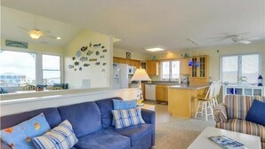 Een woonkamer met een blauwe bank, een gestreepte fauteuil en een witte salontafel. Er is een keuken met houten kasten en een raam.