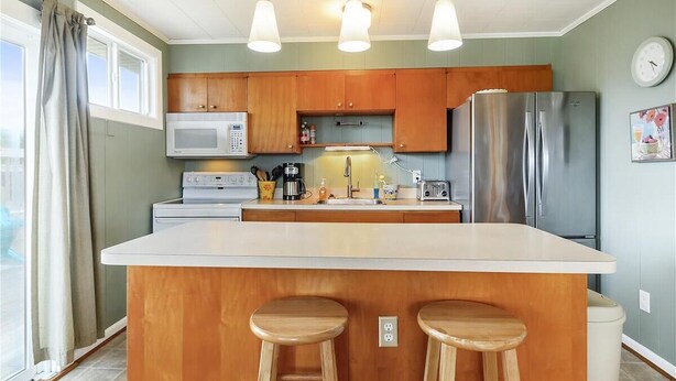 Une cuisine avec des meubles de rangement en bois, un réfrigérateur en acier inoxydable, un four à micro-ondes, une machine à café et deux tabourets en bois.