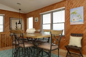 A wooden-paneled room with a round table and chairs, a painting of a beach scene, and a window with blinds. - Semi-oceanfront with a hot tub in quiet South Nags Head! (Nags Head)
