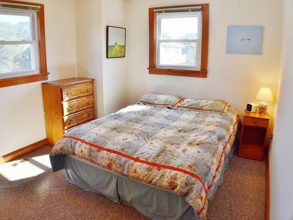 Quarto com cama, penteadeira, mesa de cabeceira e duas janelas.