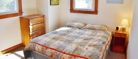 Quarto com cama, penteadeira, mesa de cabeceira e duas janelas.