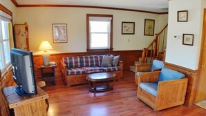 Una sala de estar con piso de madera, un sofá a cuadros, un sillón azul, una mesa de centro y un televisor sobre un mueble de madera.
