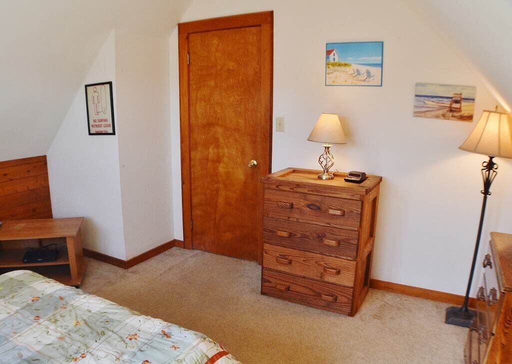 Une chambre avec une commode en bois, un lit avec une literie à motifs floraux, un banc en bois, une lampe et une porte.