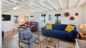 Ein geräumiges Wohnzimmer mit hoher Decke, einem blauen Sofa mit gelben Kissen, einem roten Ledersessel und einem grauen Sessel.