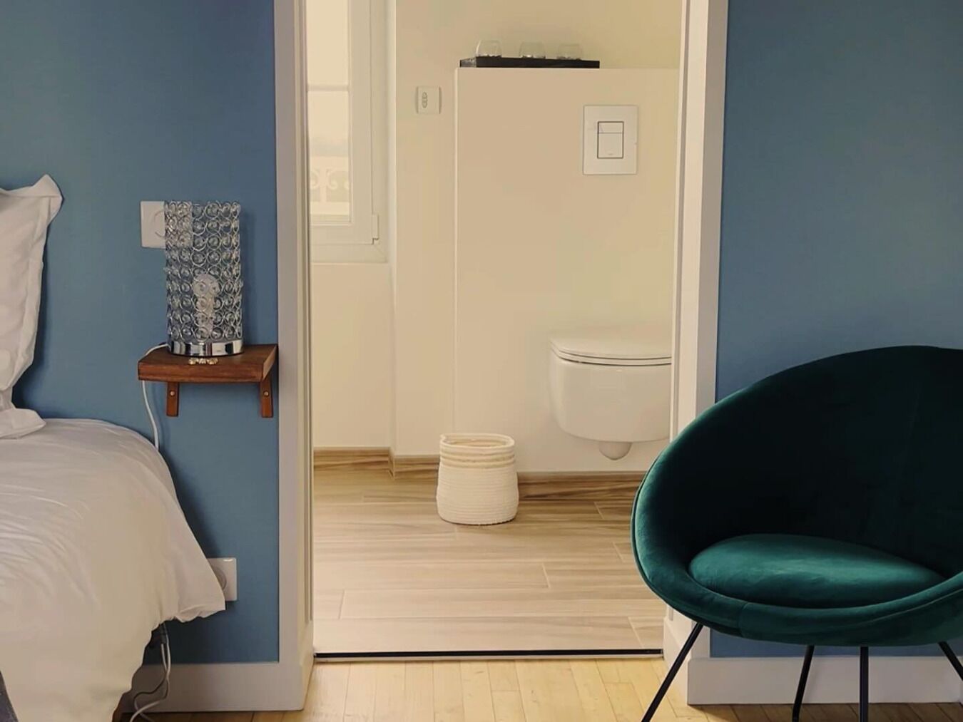 Une chambre à coucher avec un lit, une table de chevet agrémentée d’un objet décoratif, un fauteuil turquoise et une vue sur la salle de bain, où l’on aperçoit des toilettes et une douche.