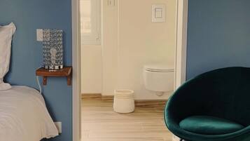 Une chambre à coucher avec un lit, une table de chevet agrémentée d’un objet décoratif, un fauteuil turquoise et une vue sur la salle de bain, où l’on aperçoit des toilettes et une douche.