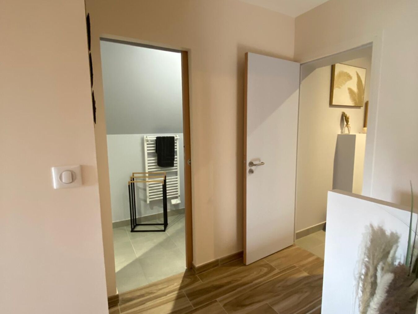 A modern hotel room with a wooden floor, a white door, and a hallway leading to a bathroom with a towel rack and a radiator.