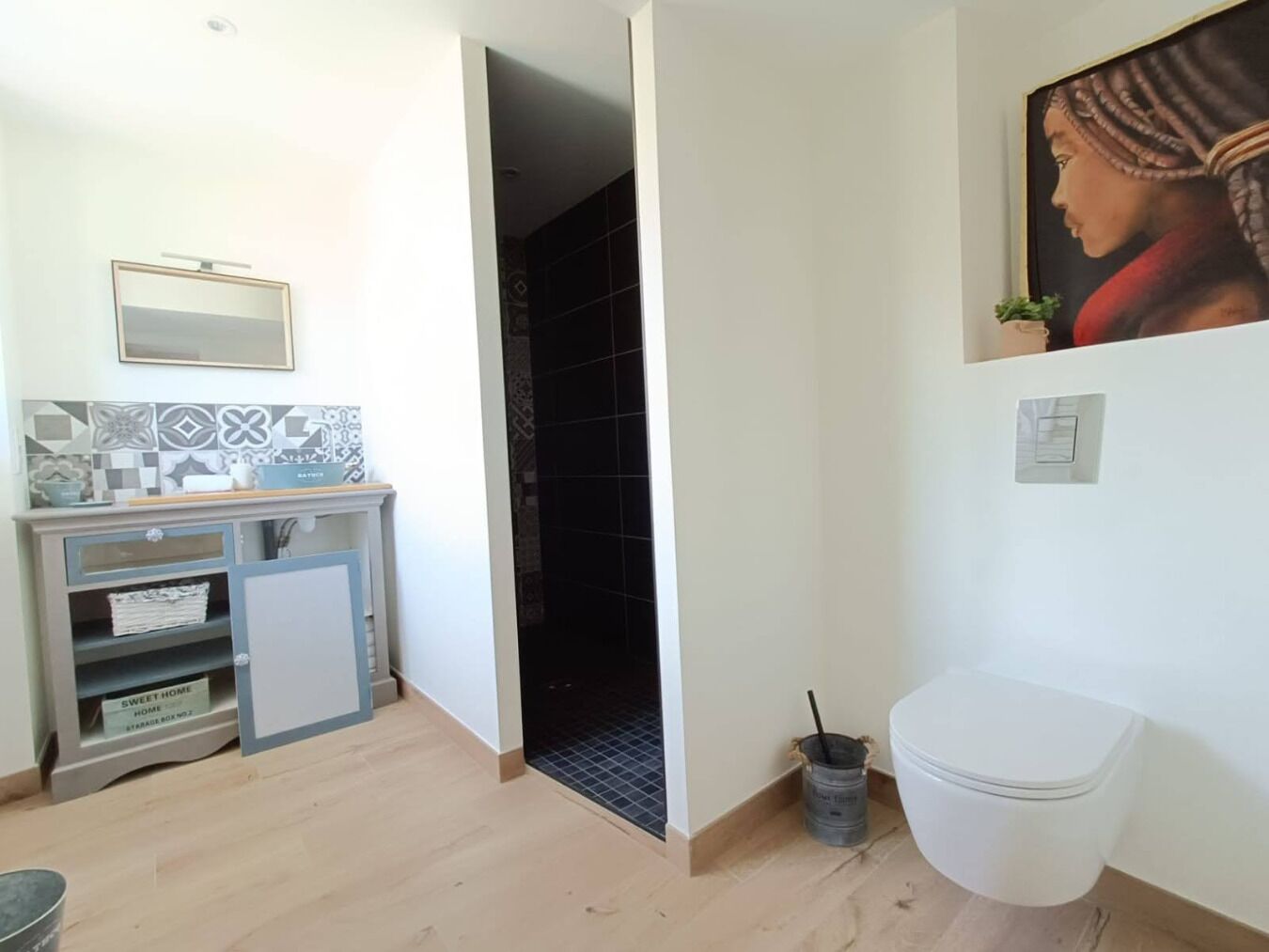A bathroom with a toilet, a sink, and a mirror. There is a tiled backsplash above the sink and a painting on the wall.