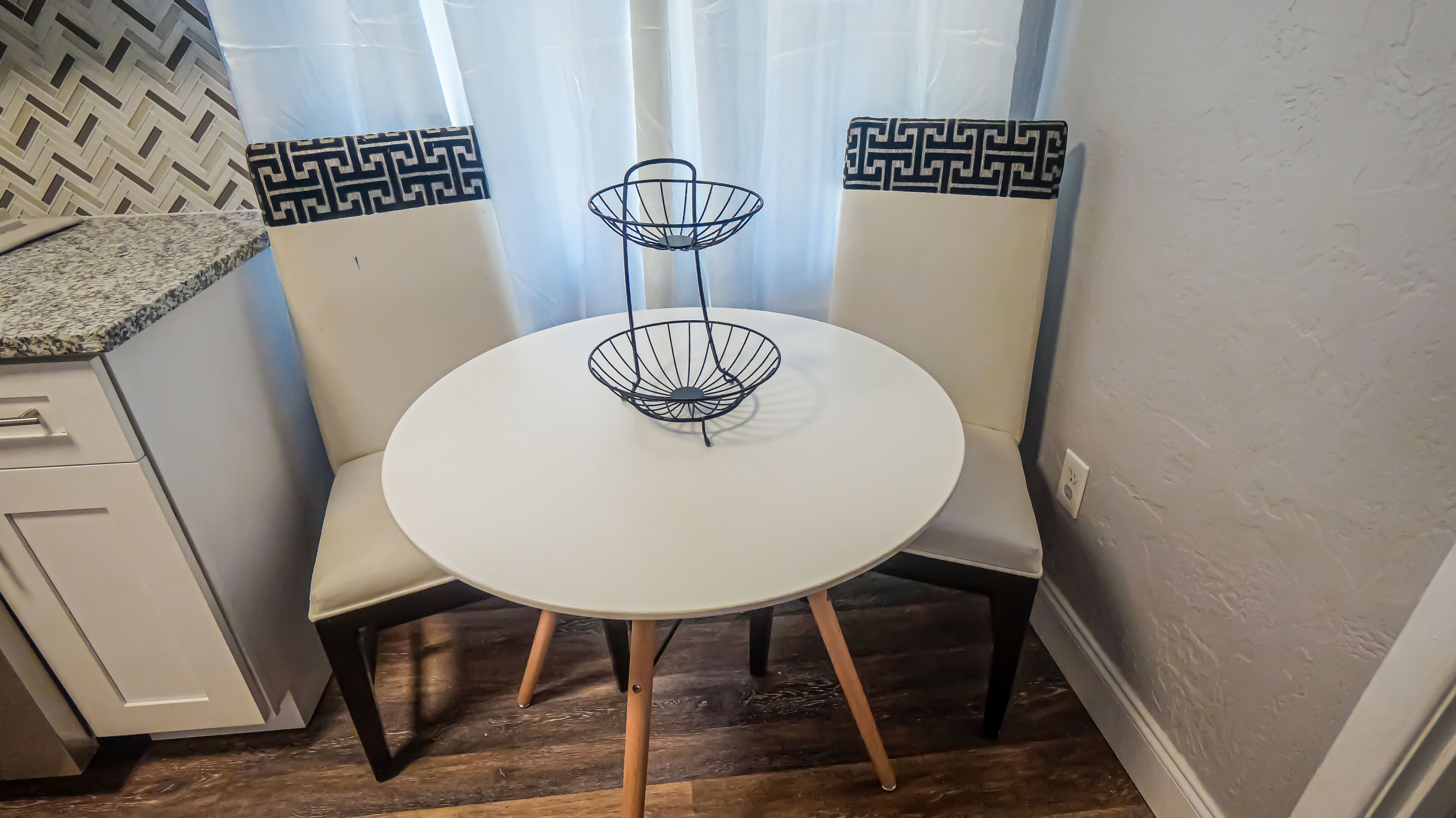 Un petit coin repas avec une table ronde blanche, deux chaises à dossier décoré et un panier en fil de fer noir posé sur la table.