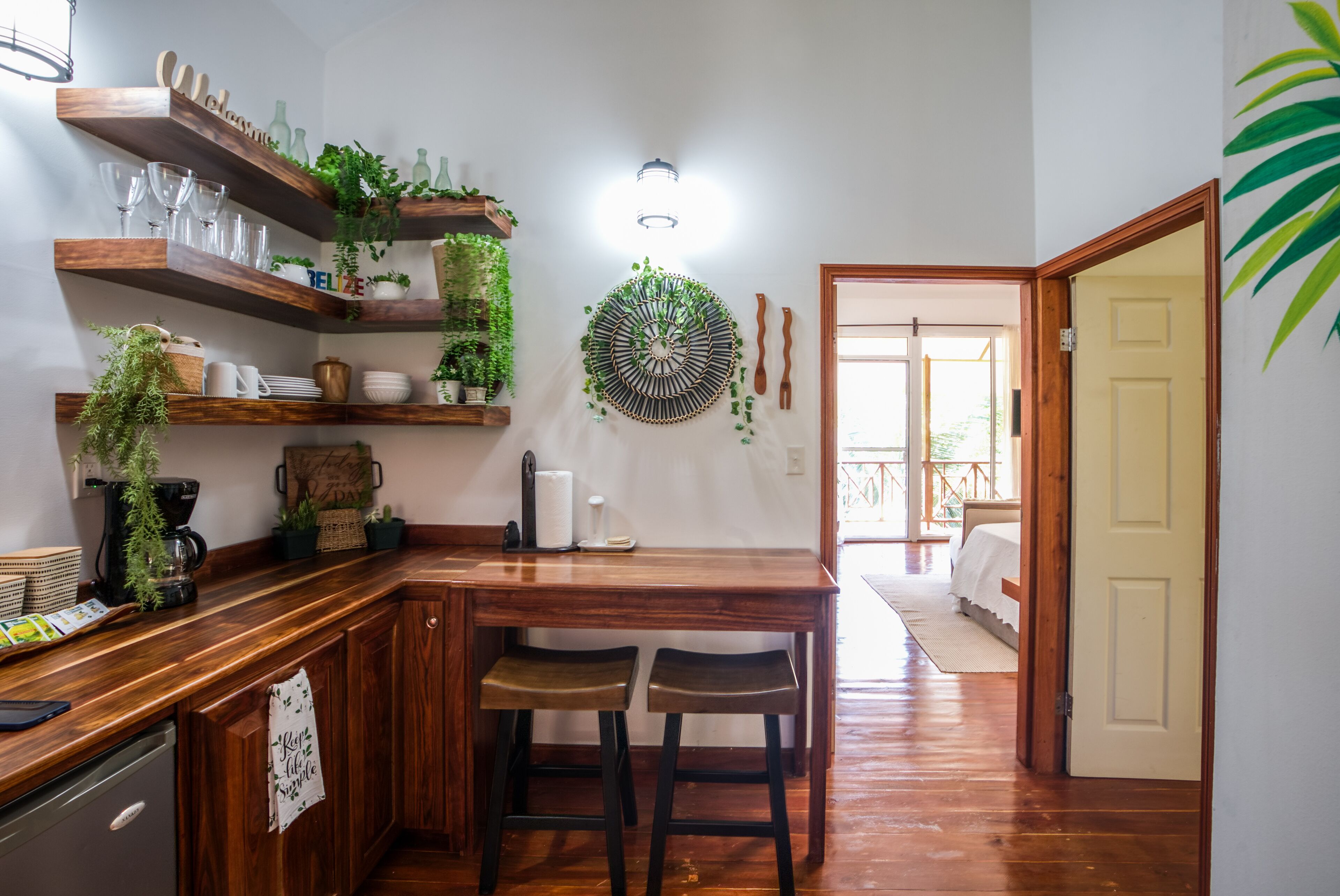 Una cucina con scaffali in legno, un tavolo da pranzo e sgabelli da bar. Ci sono piante sugli scaffali e un lampadario a parete.