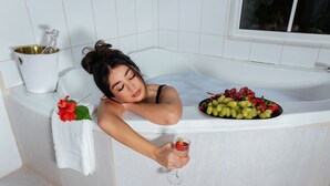 En person ligger afslappet i et badekar med et glas drik og en skål med frugt.