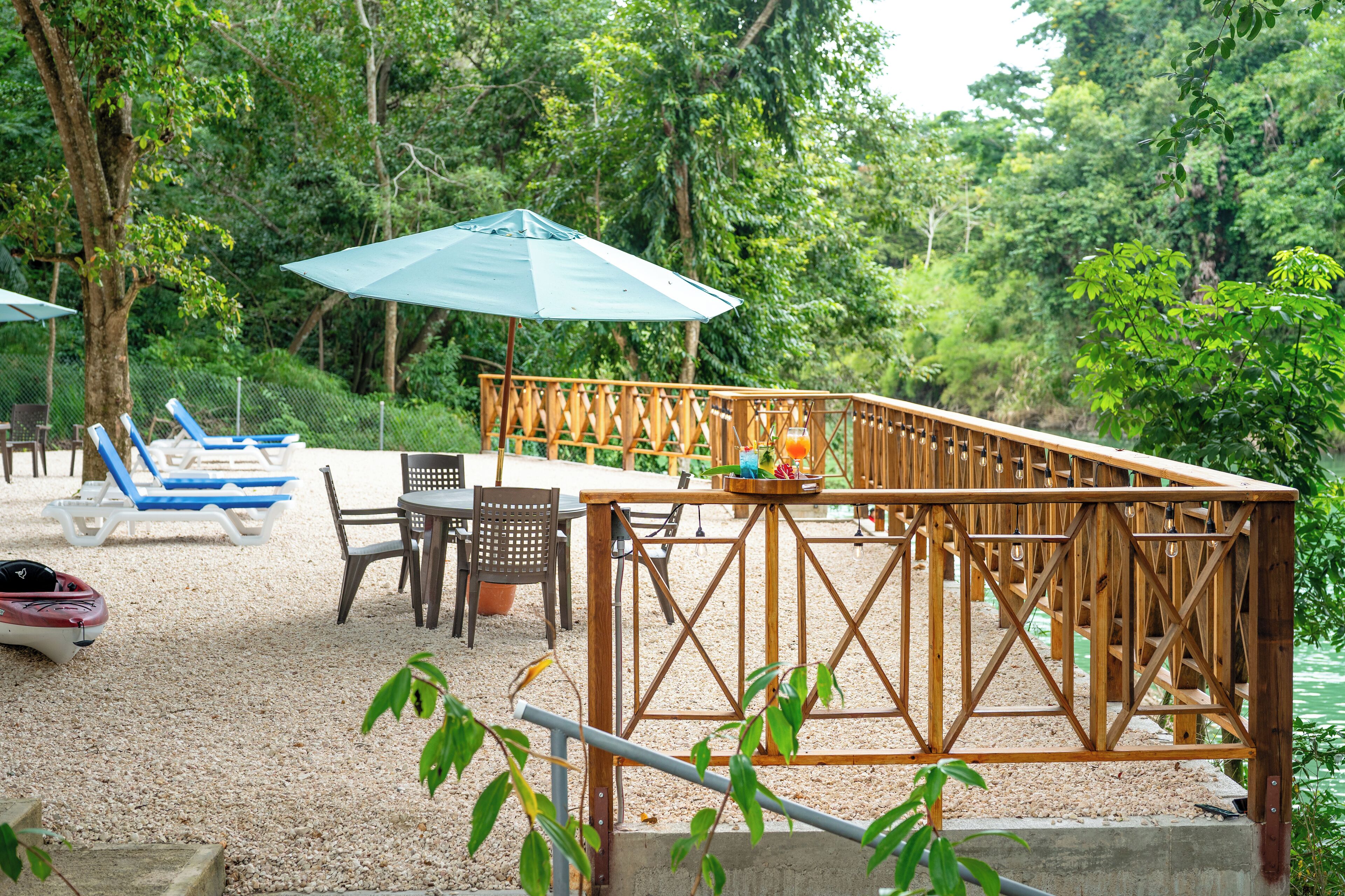 A wooden deck with a table and chairs, surrounded by blue lounge chairs and umbrellas, set against a backdrop of dense greenery.