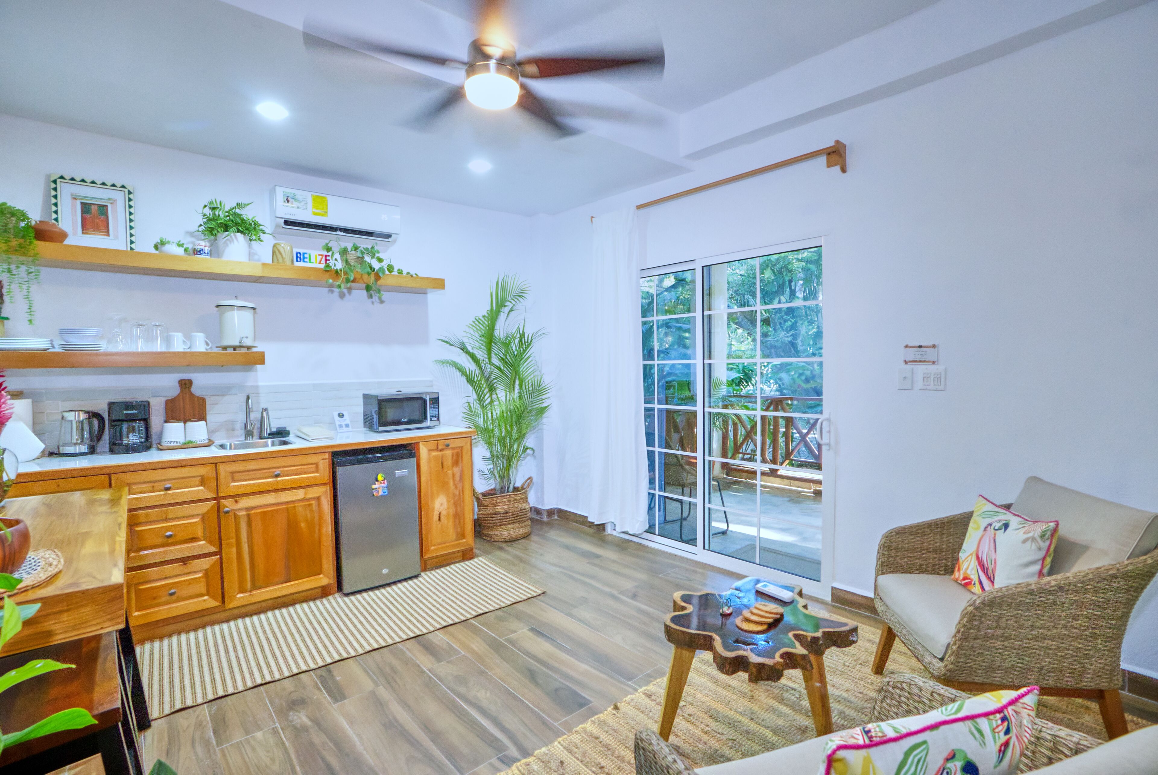 A modern kitchen with wooden cabinets, a built-in microwave, and a refrigerator. There is a dining area with a wicker chair and a small table. A large window provides a view of the outdoors.