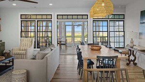 Amplia sala de estar con una gran mesa de comedor de madera, un sofá y una silla. Hay ventanas grandes con marcos negros, una lámpara colgante de ratán y vistas al exterior.