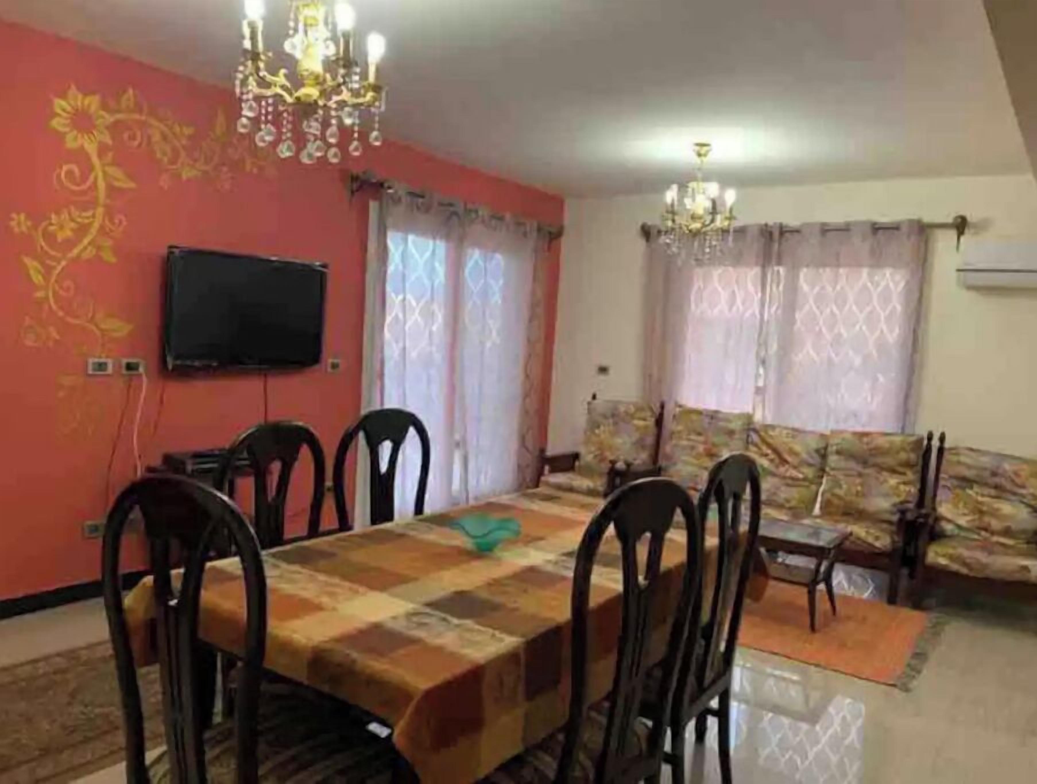 Un área de comedor con una mesa y sillas, un televisor en la pared y un sofá con cojines estampados.